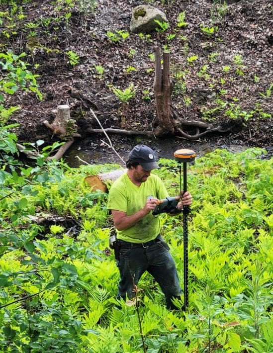 Man using surveying equipment in dense greenery.