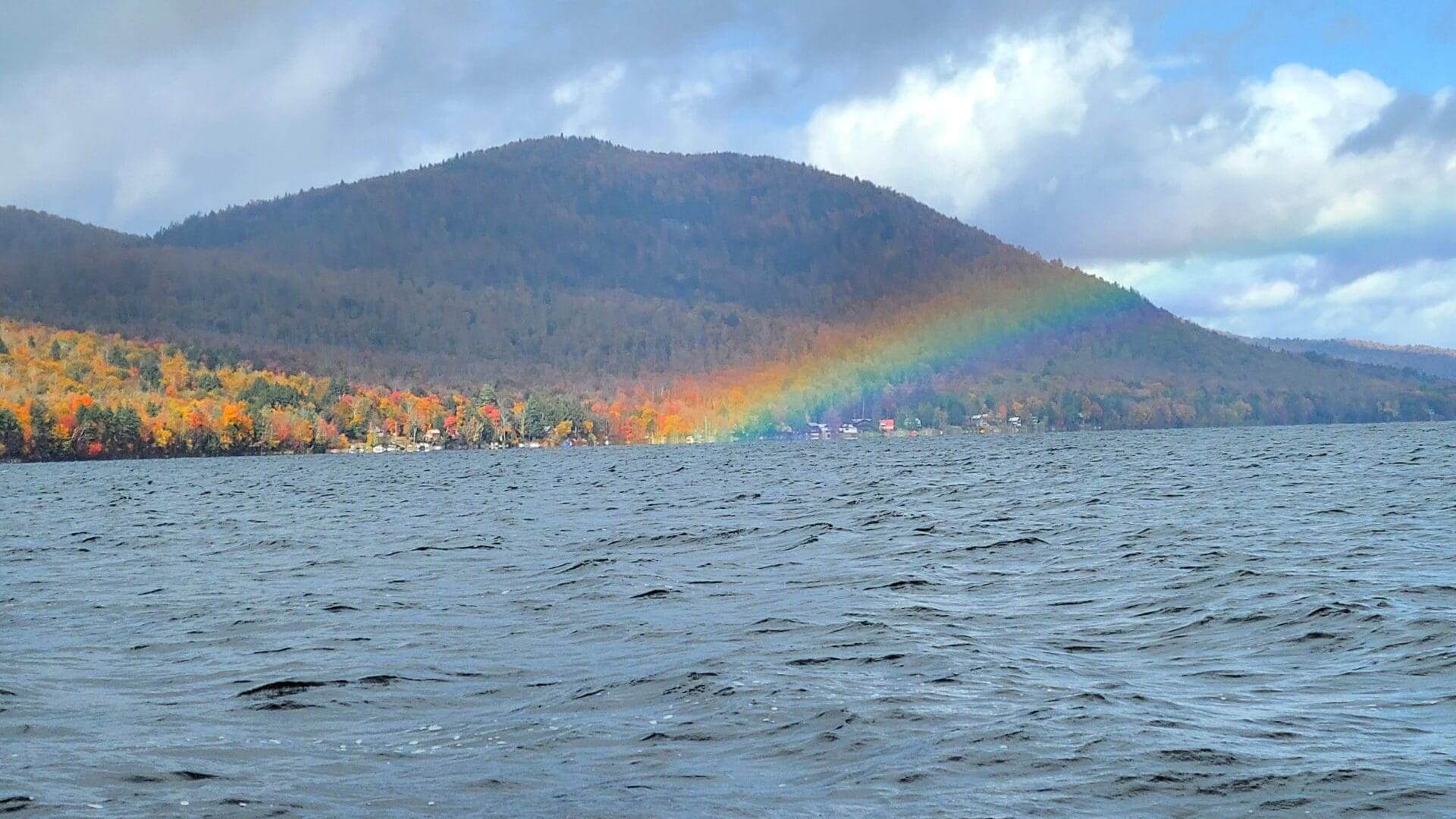 A vibrant rainbow arches over a lake with wooded hills in the background.