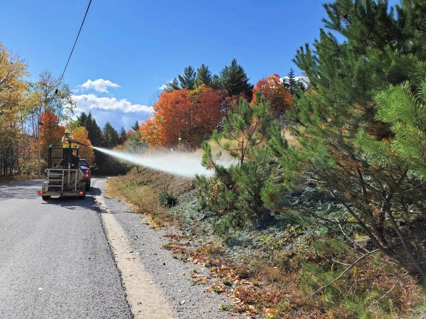 A truck sprays water along a roadside with autumn trees.